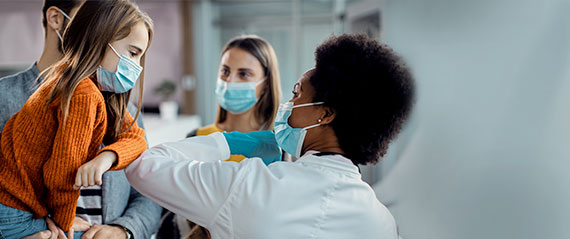 Young girl elbow bumps female doctor in a lab coat while mom looks on, all wear masks.